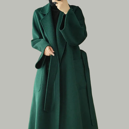 Person wearing a long green coat in a room with wooden walls and a beige chair.