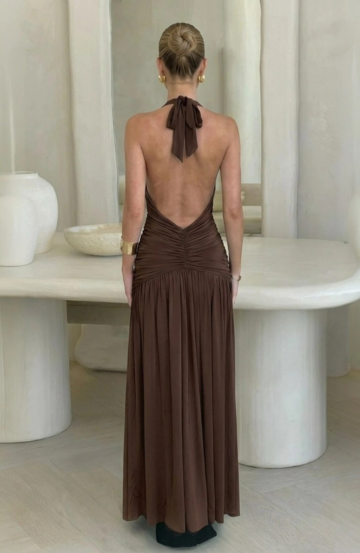 LORVENES brown ruched halter neck maxi dress with a low back and tie detail. Model wears gold earrings and bracelet.